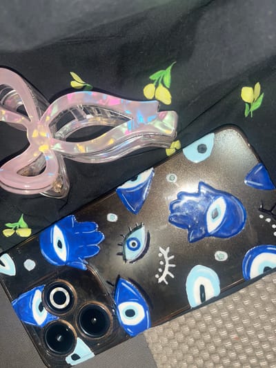 This is a top-down, close-up photograph of a black fabric with a yellow floral pattern. A transparent pink hair claw clip with an iridescent sheen is positioned on the upper left side of the frame, partially obscuring the fabric. To the right of the hair clip, a clear phone case is laid flat, decorated with blue and white evil eye symbols and hand shapes. The phone case covers the lower right portion of the image. The background fabric appears textured, and the overall lighting suggests an indoor setting. The location in Conuco Viejo, Venezuela is not discernible from the visual information in the image.