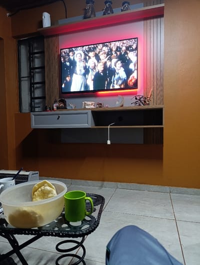A TV mounted on a wall display shows a historical event. Below, a floating shelf holds decorative items. A table in the foreground contains a bowl of food and a green mug. The floor is tiled. Part of a person wearing blue clothing is visible in the lower right corner. The lighting in the room is soft, and the walls are a warm orange color.