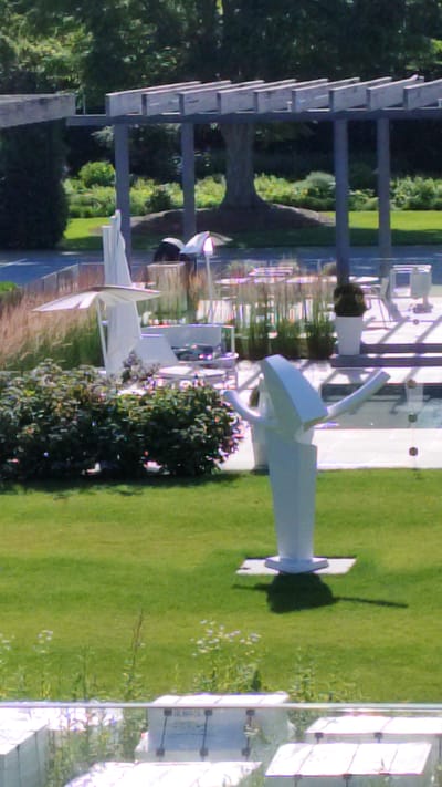 The image depicts a modern outdoor space, possibly a public area or a well-maintained garden. The scene is dominated by a manicured green lawn and lush trees, suggesting a pleasant day with good weather, likely daytime given the bright lighting.

Several contemporary white sculptures are scattered across the lawn, adding an artistic element to the environment. One prominent sculpture is a large, abstract white structure with curved, wing-like elements, casting a distinct shadow on the grass. Another sculpture, appearing to be a stylized bird or abstract wing, is also visible. These sculptures suggest a contemporary art installation or a feature of a modern architectural design.

In the background, a covered walkway or pergola structure with horizontal beams is visible, providing shade and architectural interest. Beyond this, glimpses of buildings with glass facades and outdoor seating areas can be seen, hinting at a commercial or recreational establishment, such as a museum, gallery, or a high-end resort. There are also indications of water features, possibly reflecting the surroundings.

The overall impression is one of tranquility and aesthetic appeal, with a blend of natural landscape and modern design. The lack of visible people suggests a quiet moment in this location, allowing the sculptures and the environment to be the focal points. The scene is set in Bridgehampton, USA, contributing to the context of a potentially upscale or artistic location within the United States.