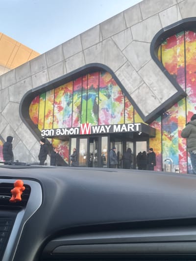 The image shows the entrance of a building, likely a store or market, taken from the perspective of someone inside a car. The building features a distinctive facade: the upper part is covered in light grey, rectangular tiles or stone, while the area around the entrance boasts a vibrant, multi-colored mural or decorative panel. This panel displays horizontal stripes of various bright colors including red, yellow, green, blue, and orange, with abstract, possibly painted, patterns overlaid.
Above the entrance, a prominent black sign with white lettering reads "W WAY MART." Below the Latin script, there is additional text written in Georgian script, which is partially visible. The entrance itself consists of multiple glass doors.
Several individuals are gathered near the entrance, appearing to be entering or exiting the building. They are dressed in warm, dark winter clothing, including jackets and hats, suggesting cool or cold weather. One person is also visible higher up on the building's facade, also in winter attire.
In the foreground, on the right side of the image, parts of a car's interior are visible, including a dark grey dashboard and an air vent. An orange, humanoid-shaped figurine is attached to one of the car's air vent slats. The sky in the upper left corner is clear and light blue, indicating that the photo was taken during the daytime under bright conditions.