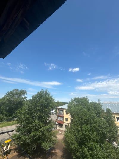 The image is a wide-angle shot taken from a high vantage point, looking up at a bright blue sky with scattered white clouds. In the foreground, a dark wooden overhang partially obscures the view. Below the sky, a dense canopy of green trees dominates the middle ground, with a small apartment building with yellow and brown siding visible between them. The building has multiple windows and balconies. Beyond the trees, a road and some other structures can be seen. The overall scene suggests an outdoor environment, possibly a residential area in a city. The date stamp indicates the image was taken on July 28, 2022, at 11:48 AM.