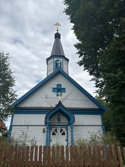 The image displays the frontal exterior of the Saint Mary Orthodox Church in Wojnowo, Poland, under an overcast sky. The church is constructed primarily of light-colored, possibly white, horizontal wooden siding accented by intricate blue decorative trim along its gables, eaves, and architectural elements. An octagonal belfry with a dark conical roof is centered at the top, crowned by a golden Orthodox cross featuring a triple-bar design. Below the belfry, the main facade presents a prominent blue square panel embedded with another golden Orthodox cross. The entrance consists of double white wooden doors with small windows, sheltered by an ornate, curvilinear blue wooden porch structure that includes a single hanging lamp. A weathered brown wooden picket fence is positioned in the foreground, with sparse green foliage visible between the fence and the building. Dense green deciduous trees frame the church on both sides and extend behind it. No individuals or active elements are present in the scene.