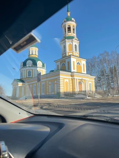 A view through a car windshield captures the Church of the Holy Trinity in Chutyr, Russia. The church is painted in yellow and white, standing prominently against a bright blue sky. Bare trees line the background. The car's interior frames the view of the church.