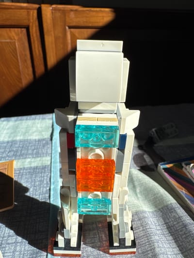 The image shows a LEGO robot standing on a blue and white checkered surface, likely a bedspread. The robot is predominantly white, with transparent blue and orange LEGO bricks forming its torso. To the left of the robot, a shadow of a person is cast on the surface. In the background, a dark wooden cabinet with vertical panels is visible, illuminated by sunlight. The scene is devoid of any indications of Sant'Angelo di Piove di Sacco, Italy, and no people are present in the frame.