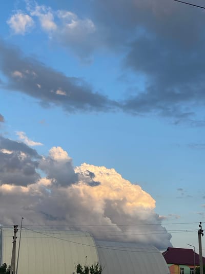 This is a vertical, eye-level, outdoor photograph taken in the city of Krasnokutsk, Ukraine. The image features a dramatic sky filled with clouds, illuminated by the warm glow of sunset. The sky is a vibrant blue, transitioning to darker, ominous gray clouds in the upper portions and to the left, suggesting an approaching storm or the remnants of one. A large, fluffy cumulus cloud dominates the center of the frame, catching the sunset light and appearing almost golden. Below the sky, a curved, light-colored building with vertical corrugated siding is visible, possibly a warehouse or an arena. A dark-colored roof of a house with a visible window is seen to the right of the large building. Overhead, power lines and utility poles stretch across the frame, indicating the presence of infrastructure. The overall scene captures a moment of natural beauty and impending weather change.