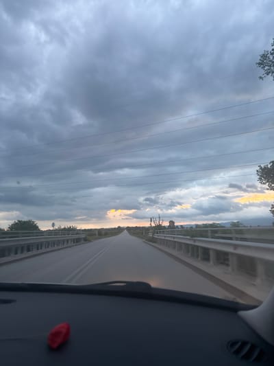 The image shows a view from inside a vehicle driving on a road. The sky is predominantly cloudy with patches of lighter clouds, possibly indicating sunset. Power lines run parallel to the road. The road has a guardrail. There is a small, red object visible on the dashboard. The photo was taken in Lesnovo, Bulgaria.