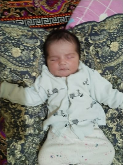 A newborn baby is lying on its back, asleep, on a patterned blanket. The baby is wearing a light blue onesie with a pattern of scooters and small text. The blanket has a dark blue base with a repeating gold and white floral pattern. Behind the baby, to the left, is a section of a colorful, ornate fabric with purple, orange, and black patterns. To the right of the baby is a pink and white checkered pillow. The lighting is moderate, suggesting an indoor setting. The image appears to be a photograph taken in a home environment, possibly in the city of Jizzakh, Uzbekistan, given the provided location context.