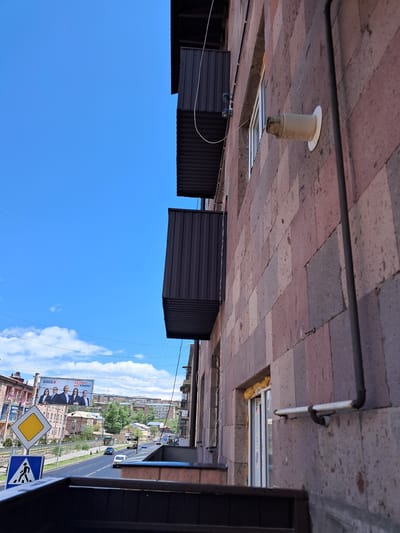 The image, taken from an elevated perspective, captures a view looking down onto a street scene, with an overhead structure dominating the foreground. The photograph appears to be taken during the daytime under clear, sunny weather, indicated by the bright blue sky with scattered white clouds.
In the immediate foreground, a tiled ceiling, composed of reddish-brown and gray rectangular pavers, stretches across the top of the frame. Attached to this ceiling are dark gray and white pipes, a round, beige recessed light fixture or vent, and a rectangular recessed light fixture with visible "citilux" branding. Exposed wiring also runs along the underside of this structure.
Below this overhead structure, a series of dark brown, corrugated metal awnings or shelters are visible, partially obscuring the middle ground. Beyond these, the open blue sky fills a significant portion of the image.
The background reveals a bustling street scene. A multi-lane road with cars and buses is visible, flanked by buildings of varying architectural styles and lush green trees. Power lines crisscross the sky above the street. Prominently positioned on the right side of the road are two road signs: a yellow diamond-shaped sign indicating "Priority Road" and a blue square sign depicting a pedestrian, signifying a "Pedestrian Crossing."
A large billboard stands out on the left side of the street, featuring a group of people and text. Visible Armenian text includes "17", "ԲԱՐԵԿԵՆ" (Bareken), "ՀԱՄԱՇՐՋԱՆ" (Hamashrjan), and "ԹԱՐՄԱՑՈՒՄ" (Tarmatsum), along with other unreadable text. The overall scene suggests an urban environment in Gavar, Armenia.