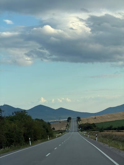 The image depicts an empty asphalt road stretching into the distance towards rolling hills and mountains under a cloudy sky. The road is lined with white and red reflective markers. The surrounding landscape consists of fields, vegetation, and scattered trees. In the background, a prominent peak with what appears to be a communication tower is visible. The scene is devoid of any people or vehicles, suggesting a quiet, rural setting. The weather appears overcast but not actively raining. This location is identified as Luhyňa, Slovakia.