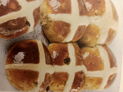 The image shows a close-up, overhead view of several hot cross buns arranged in a cluster. The buns are golden brown with a white cross piped on top, dusted with a light coating of white powder, likely flour or powdered sugar. Some raisins or other dried fruit are visible embedded within the buns. The background is a textured, neutral surface. There are no people, actions, interactions, or environmental details related to the city of Rumaithiya, Kuwait, or any specific location present in the image.