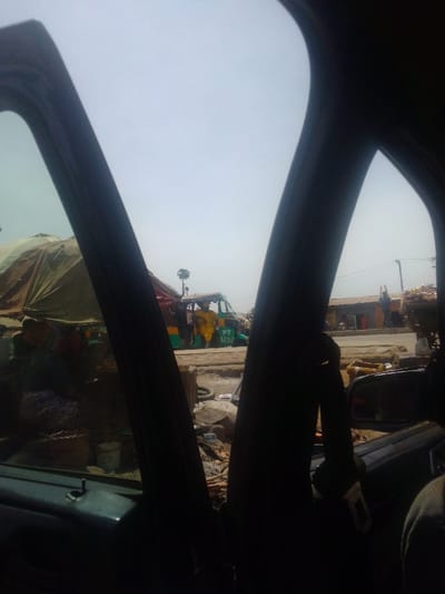 The scene depicts an outdoor street in Mista Ali, Nigeria, viewed from inside a vehicle, framed by dark interior pillars and door components. The immediate foreground reveals unpaved ground littered with debris. To the left, a makeshift market stall is covered by a large, light brown tarp, with goods and containers visible beneath it, and a person partially discernible, seated nearby. Centrally in the midground, a green and yellow motorized tricycle, marked with "PT 1638," is stationary. A person wearing a yellow garment stands next to the tricycle. A solitary palm tree rises behind the tricycle against a bright, hazy sky. To the right, along a dirt road, several single-story structures with corrugated roofs line the thoroughfare. Multiple individuals are present near these buildings, one figure in the distance has arms raised overhead. Overhead power lines are visible in the background.
