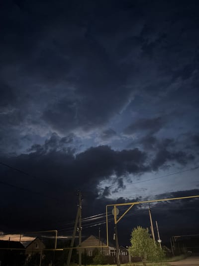 The media file depicts a night scene with a dramatic sky filled with dark, ominous storm clouds. The clouds dominate the left side of the image, appearing dense and turbulent, with hints of lighter sky peeking through in some areas. On the right side of the frame, a utility pole with numerous wires stretches upwards. A prominent yellow pipe runs along the pole, possibly indicating a gas line.

In the background, on the far right, there are glimpses of residential buildings with lights on, suggesting a rural or suburban setting. A tree with green foliage is visible, its leaves illuminated by an unseen light source. The overall atmosphere is moody and charged, likely due to the approaching storm. The time of day is clearly night, as indicated by the darkness and the presence of artificial lights. There is no visible text in the media file. The setting appears to be Nikolaevka, Russia, as per the provided context. The image conveys a sense of anticipation and perhaps unease due to the impending weather.