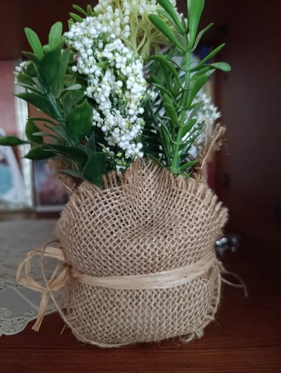 The image shows a close-up of a small artificial floral arrangement in a burlap pot. The pot is wrapped in a coarse woven burlap fabric, secured with a thin, light-colored ribbon tied in a bow on the left side. The burlap has frayed edges. The floral arrangement consists of green faux foliage and clusters of small white faux flowers, with some yellow accents visible. In the background, on a wooden surface, is a white lace doily to the left and a dark brown shelf or cabinet to the right. Out of focus in the background are framed pictures. The scene appears to be indoors, possibly on a shelf in a home or shop. There are no people visible in the image. The location is not specified as being in the city of Criuleni, Moldova.