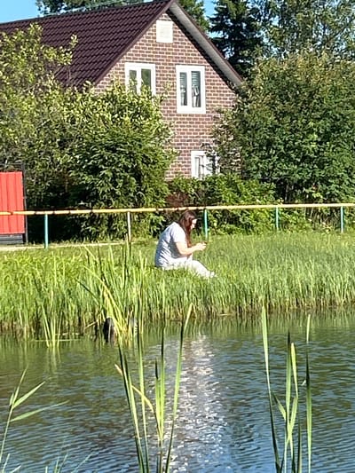 A woman is sitting in the tall green grass on the bank of a body of water. She is wearing a light-colored, patterned shirt and pants and appears to be looking at her phone. Behind her, there is a yellow railing with green posts, and further back is a brick house with a dark roof and trees. The water in front of her is rippled, reflecting the sunlight. The scene appears to be in a suburban or rural area.