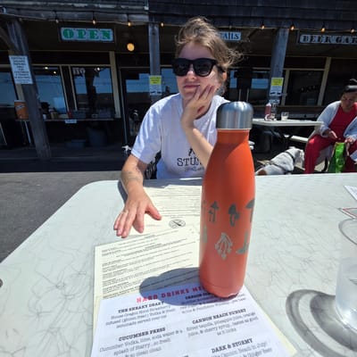 In Cannon Beach, Oregon, at an outdoor seating area adjacent to a building with signage indicating "OPEN" and "BEER GARD," a woman wearing sunglasses and a white t-shirt with a graphic print sits at a table. She is resting her chin on her hand, looking slightly to the side. In front of her on the table is an orange water bottle with a metallic lid, decorated with mushroom graphics. A menu is also visible on the table. Further back, a bulldog on a leash is visible, and a woman in a red dress is seated. The environment appears to be a sunny day, with shadows cast on the table.