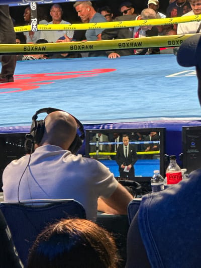The image depicts an indoor boxing event, likely a live production or broadcast setup. The exact venue within Winchester, United States, is not discernible from the image.
In the immediate foreground, from a rear perspective, a bald male wearing black over-ear headphones and a light grey short-sleeved polo shirt is seated at a workstation. He faces a flat-screen monitor that displays a live feed of a male announcer. The announcer, dressed in a dark suit, white shirt, and dark tie, holds papers and stands within a boxing ring. The monitor shows a "4K" designation in its upper right corner. To the right of the monitor, on the workstation, a plastic water bottle and a red Coca-Cola bottle are visible, along with a dark, vertical piece of equipment.
The physical boxing ring, partially visible beyond the workstation, features a blue canvas with red markings. It is enclosed by yellow ropes bearing the black text "BOXRAW" and a white rope displaying "RING" in black text. Behind these ropes, several individuals are seated, observing the event. Notable figures among the spectators include a man with short gray hair wearing a light blue t-shirt and a white earbud in his left ear, and another man with short, lighter-colored hair in a white shirt, holding a blue object. A woman with dark skin and short dark hair is also visible, smiling. Partial views of other individuals' heads and shoulders appear in both the extreme foreground and the background.