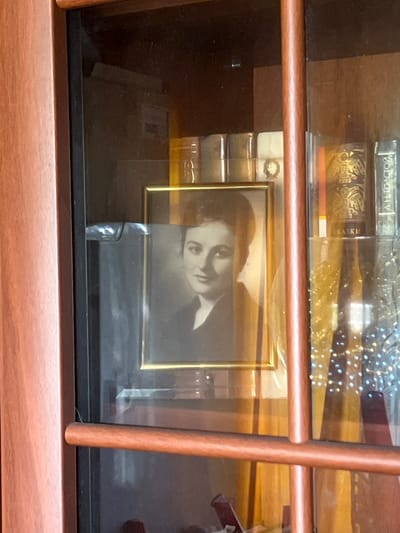 A framed black and white photograph of a woman with dark hair and a soft smile is displayed inside a glass-fronted wooden cabinet. The photograph is gold-framed and positioned centrally within the cabinet. Behind the photograph, several books with decorative spines are visible on shelves. The cabinet doors have wooden trim, and vertical and horizontal wooden dividers are present. The lighting suggests an indoor environment, possibly a home or a shop. The image is a close-up shot, capturing the details of the photograph and its surroundings.