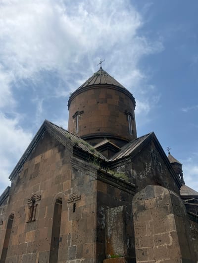 The image shows a low-angle view of Saghmosavank Monastery in Artashavan, Armenia. The architecture is dominated by dark stone construction against a backdrop of a cloudy blue sky. The dome and roofline of the religious structure are visible, featuring a cross at the apex. Patches of vegetation are growing on the structure's surface.