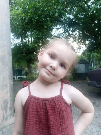 This is a portrait of a young girl with light brown hair, styled in pigtails. She is wearing a reddish-brown sleeveless dress with thin straps and a textured fabric. Her head is tilted to her left, and she is looking directly at the camera with a slight smile. The background is a soft blur of green foliage from trees, suggesting an outdoor setting. The lighting appears to be natural, and given the foliage, it's likely daytime. In the blurred background, there are hints of structures, possibly a shed or fence, and a dark-colored vehicle on the right side. A rough, light-colored wall or pillar is visible on the left side of the frame. The overall mood is casual and candid, capturing a moment of the girl's playful expression. The scene is set in Yampil, Ukraine. There is no visible text in the image.