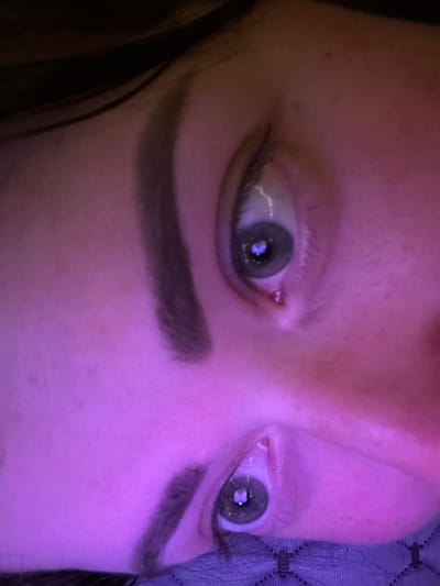 An extreme close-up, low-angle, over-the-shoulder shot shows the face of an individual. The lighting is predominantly purple, with some pink hues. The subject's left eye is visible, with a green iris and what appears to be a reflection of a heart shape in the pupil. A trace of red is present on the lower eyelid. The subject's eyebrows are dark and appear to be filled in. The right eye is partially visible, also with a green iris. The skin has some blemishes. The background is dark and out of focus, with hints of texture that may be fabric. The location is Lynnwood, United States.