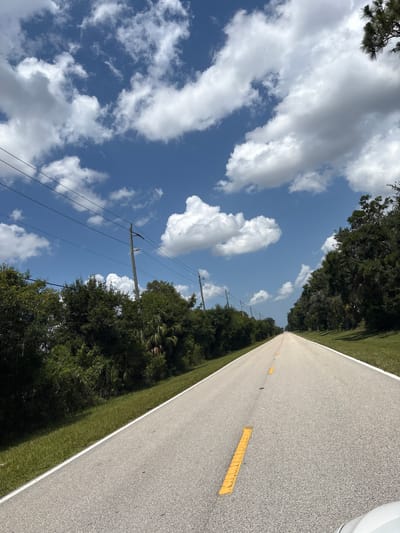 The image shows a long, straight asphalt road with a white line on the left edge and yellow dashed lines in the center. The road is lined with green grass on both sides, which transitions into lush green trees and vegetation. Utility poles with power lines are visible on the left side of the road. The sky is a vibrant blue with scattered white, puffy cumulus clouds. The perspective is from a low angle, looking down the road as if from a vehicle. There are no people or animals visible in the scene. The lighting suggests it is daytime, with the sun casting shadows from the trees. The environment appears to be rural or suburban, with undeveloped land and natural vegetation. There is no specific indication of the city of Bradenton, United States of America.