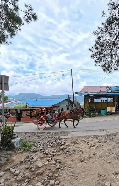 A horse-drawn carriage, carrying several people, travels on a paved road in front of shops. A brown horse pulls the carriage, which is decorated with colorful fabric. The carriage has spoked wheels and a canopy. In the background, buildings with red and blue roofs are visible, along with a distant mountain range. The sky is cloudy. The foreground of the image is a rocky, unpaved area. The scene is located in Gundaling 2, Indonesia.