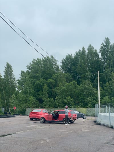 The image shows an outdoor scene in Ковалёво, Russia. The foreground is a cracked asphalt surface. In the middle ground, two red cars and a dark-colored car are parked. One red car is open, with its doors and trunk removed. A person wearing a pink helmet, a gray sweatshirt, and dark pants is standing next to the open red car, facing the camera. In the background, a dense line of green trees forms a backdrop under a gray, overcast sky. Power lines are visible stretching across the sky. To the right, a section of a metal fence with a white barrier is present. A red and white stop sign is visible to the left of the red cars.