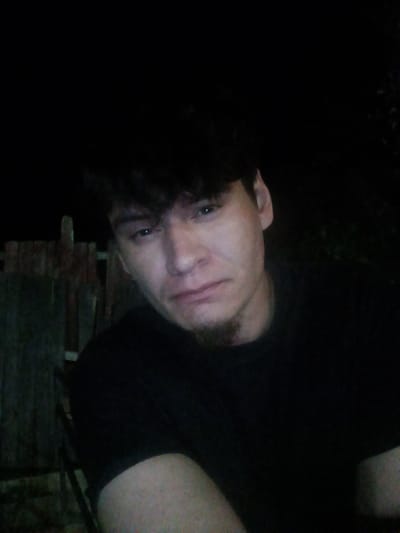The image depicts a person with dark hair and a small goatee, wearing a black shirt. The person appears to be outdoors at night. A wooden fence is visible in the background. Based on the prompt requesting the location in Fort Smith, United States of America, the image may have been taken there. The dark and obscured background makes it difficult to determine the exact location.