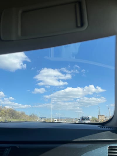 The image is taken from inside a vehicle, looking upwards through a sunroof. The sky is visible, displaying a bright blue hue punctuated by fluffy white clouds. In the distance, through the windshield, there is a highway. Cars and other vehicles are present on the highway. Utility poles and industrial buildings can be seen in the background. Some greenery is visible along the roadside.
The location is Balgarchevo, Bulgaria. The image shows a roadside scene on a bright, sunny day. The time of year is spring, given the tree foliage.