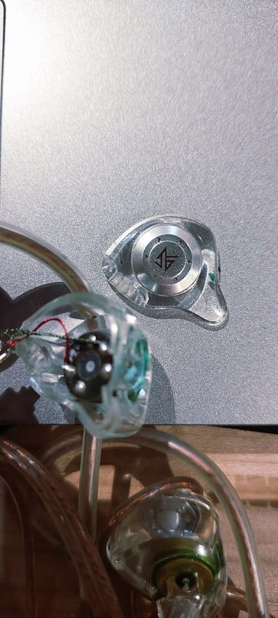 Two transparent in-ear monitors (IEMs) are displayed on a textured, silver surface. One IEM, positioned to the left, features a circular metallic plate with a black logo consisting of two opposing arrows. The other IEM, to the right and slightly out of focus, reveals internal circuitry including red and green wires and what appears to be a small speaker driver. Clear tubes, likely part of the IEMs or connected audio equipment, curve around the objects and extend out of frame. The background to the right is a warm, wooden texture, and the lighting suggests an indoor setting. The overall impression is of electronic audio equipment. No people are visible, and no specific time of day or weather conditions can be inferred. There is no discernible text other than the logo on the left IEM. The location context of Pagedangan, Indonesia, does not provide further visual cues within the image.