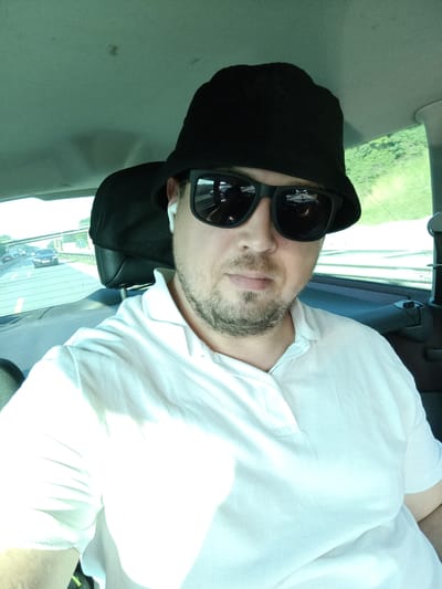 The media file shows a selfie of a person from the chest up, taken inside a vehicle. The person is wearing a white polo shirt, a black bucket hat, and dark sunglasses. They also have white wireless earbuds in their ears and a light beard. The background visible through the car window suggests it is daytime, with trees and other vehicles on a road. The setting appears to be a highway, as indicated by the lanes and cars. The overall impression is of someone casually traveling by car. There is no visible text in the image.