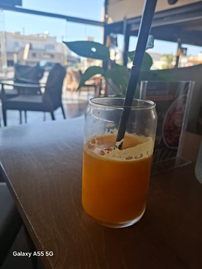 The image shows a glass of orange juice with a black straw, placed on a wooden table. In the background, visible through a window, is a cityscape with buildings and vegetation, suggesting an urban environment in Heraklion Municipal Unit, Greece. The lighting indicates it is daytime. A menu or advertising material is partially visible behind the glass. The image was captured using a Galaxy A55 5G phone.