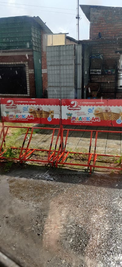 The image depicts an outdoor scene in Shelestove, Ukraine, characterized by overcast skies and visible raindrops, indicating inclement weather. In the foreground, a bright red bicycle rack stands on a wet paved surface, with a puddle reflecting the surrounding environment. Behind the bicycle rack, a banner advertising ice cream is displayed. The banner features images of ice cream cones and text in Ukrainian. Beyond the banner, buildings with brick and corrugated metal facades are visible, along with a large grey metal gate. The overall atmosphere is damp and subdued, consistent with a rainy day in an urban or semi-urban setting.