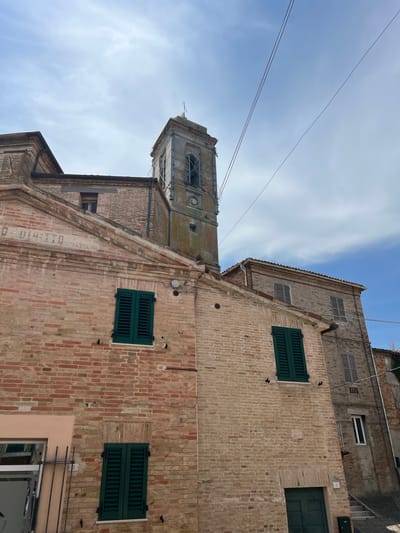 This is an eye-level, outdoor, medium shot of a weathered brick building with a bell tower in the background, located in Barbara, Italy. The sky is partly cloudy. The building in the foreground has a red brick facade with green shutters on its windows. A faded inscription is visible on the upper facade. The bell tower, also made of brick, has visible architectural details and a cross at its apex. Telephone wires stretch across the sky above the buildings. The scene is characteristic of an old Italian town.