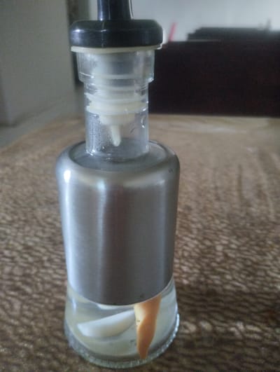 The image shows a close-up of a clear glass bottle with a stainless steel collar, topped with a black dispenser. Inside the bottle, there is a light yellow liquid with two white objects and a piece of orange, peel-like material submerged in it. The bottle rests on a textured brown surface, possibly a tablecloth or rug. In the background, a dark brown piece of furniture is visible, and further back, a blurry light-colored wall. The scene is indoors, and the lighting is diffused, suggesting an ambient light source. There is no indication of the city of Gulbakhor, Uzbekistan, or any specific location.