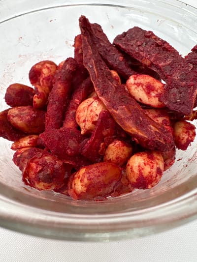 A clear glass bowl contains a mixture of chili-coated snacks. The contents consist of shelled, roasted peanuts and irregularly shaped, dark red pieces, consistent with chili-dusted dried fruit or vegetable strips. The red seasoning fully coats the surfaces of both snack types, with visible particulate residue on the interior glass surface. The pieces of dried fruit or vegetable vary in form, including elongated strips and flatter, broader segments, all exhibiting a textured, reddish-brown appearance. The snacks are loosely piled within the bowl. The bowl rests on an indistinct, light-colored surface, which is out of focus. No individuals are present, and no actions are depicted. The depicted snack items are typical of those found in Chamilpa, Mexico.