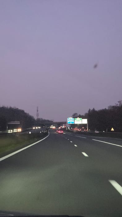 The image presents a forward-facing view from a vehicle on a multi-lane highway at twilight or night. The scene is identified as being near Briis-sous-Forges, France, confirmed by overhead gantry signs on the right displaying "A 10 E50", "D 19", and "Briis-sous-Forges". The sky is a uniform, muted purplish-gray with a dark speck in the upper right quadrant. Dense foliage lines both sides of the highway, with a prominent communication tower visible on the left side, beyond the trees.

The asphalt roadway features multiple lanes marked by white dashed lines and a solid white line on the left shoulder, partially illuminated by the foreground vehicle's headlights. Moderate vehicular traffic is present in both directions, indicated by numerous illuminated white/yellow headlights and red taillights. An overpass bridge spans the highway in the mid-distance. A yellow triangular warning sign is visible on the right shoulder. No individuals are discernible within the frame.