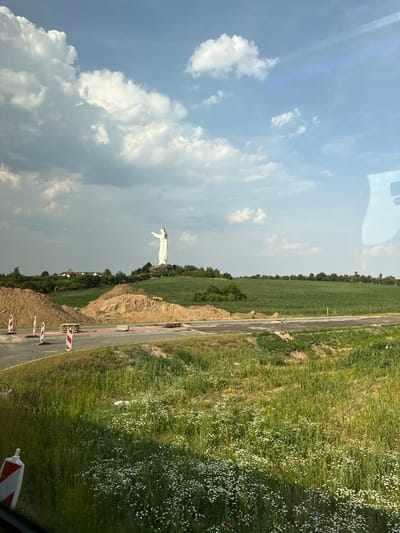 The image shows a large white statue of Christ the King on a hill in Świebodzin, Poland. The statue is visible in the distance, with its arms outstretched. In the foreground, there is a construction site with piles of dirt, traffic cones, and unfinished roadwork. The surrounding landscape is green, with fields and trees under a partly cloudy sky. The perspective suggests the photo was taken from a vehicle, possibly a bus or car, due to the reflection on the right side of the image.