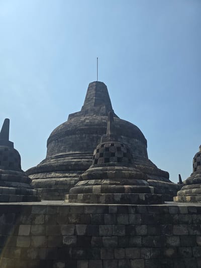 The image shows a close-up, upward-angled view of the Borobudur Temple in Desa Borobudur, Indonesia. The structure is made of dark gray stone blocks and exhibits layered, stupa-like architecture. The top of the temple features a thin metal rod. The sky is a clear, light blue.