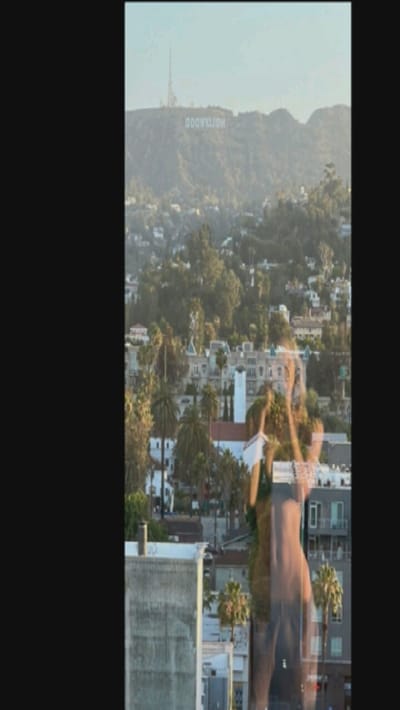 A view of the Hollywood sign sits atop a mountain, with a city and trees below. A partially transparent figure, seemingly of a woman, is overlaid or reflected in the foreground. Buildings and palm trees fill the lower portion of the image, suggesting an urban landscape. The scene is bathed in a soft, natural light, and the overall composition creates a layered effect.