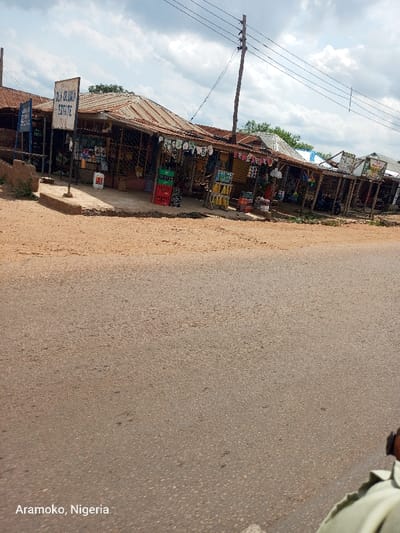 The image shows a row of shops and stalls lining a paved road in Aramoko, Nigeria. The shops are primarily constructed of wood and metal, with a sign above the first shop reading "OLA-OROKUN ESTATE". The shops appear to sell various goods including soft drinks, household items, and clothing. Some of the goods are displayed outside the shops. There is a dirt shoulder on the left side of the road, and power lines run overhead. The road appears wet, with some rain puddles visible. The image was taken from a perspective looking down the road towards the shops. The sky is partially cloudy and the weather appears to be overcast. There is a single person partially visible in the bottom right corner of the image, dressed in a green shirt, facing away from the camera. They appear to be walking on the road. The image is of a typical street scene in a developing country, with a mixture of traditional and modern elements.