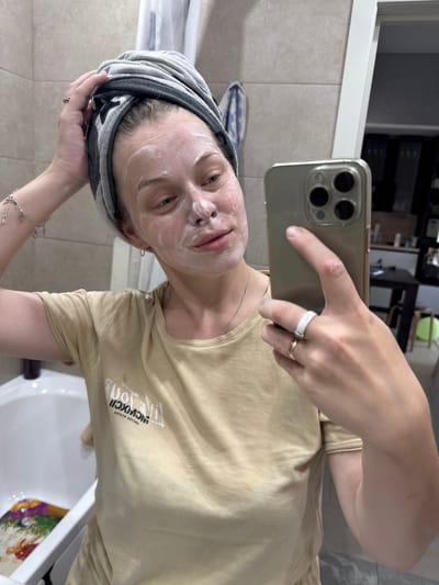 The image shows a young woman with a grey and black towel wrapped around her head and a white face mask applied to her face. She is taking a selfie in a bathroom with beige tiled walls. The woman is wearing a light brown t-shirt with white text that appears to be in Roman numerals, possibly spelling "MCMXCII" and "TITOU" or similar, and another line of text below that reads "ETATE 29TIHU". She is also wearing a silver ring on her left index finger and a delicate silver bracelet on her left wrist. The lighting suggests it is daytime, possibly indoors with artificial light contributing. The overall mood appears relaxed and focused on skincare. The background shows a portion of a mirror and what looks like shelves in the upper left corner, and a white sink and a colorful object in the lower right corner. The setting appears to be a domestic bathroom.