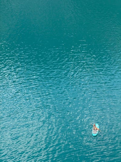 An aerial, medium-distance shot captures a person on a paddleboard in the turquoise waters of Lake Achensee, Pertisau, Austria. The person, wearing a bikini, is seated on the light blue paddleboard, holding a paddle. The water's surface is rippled, reflecting sunlight and creating patterns of light and shadow. The vast expanse of the lake dominates the frame, with the shore or any other landmass not visible.