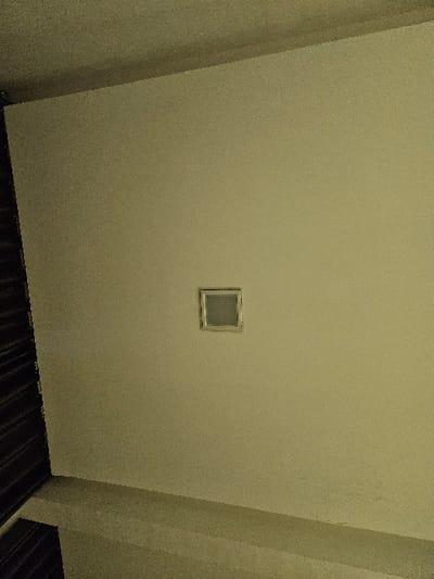 This image captures an interior architectural scene within a structure located in Padajaya, Indonesia. The dominant feature is a light-colored, likely beige or off-white, textured wall occupying the majority of the frame. Centrally embedded in the upper half of this wall is a small, square, recessed fixture, possibly a ventilation grate or a flush light, with a lighter-toned or metallic surround. The ceiling, matching the wall's color and texture, is visible along the top edge, forming an upper corner with the main wall. A dark, vertically striped object, suggestive of a closed window blind or slatted partition, is partially visible along the extreme left edge of the frame. The bottom portion of the image shows a continuous light-colored surface, presumed to be a floor or a low base wall, angled from the lower right towards the middle left. No individuals are present. The lighting is subdued, resulting in a somewhat grainy image quality.