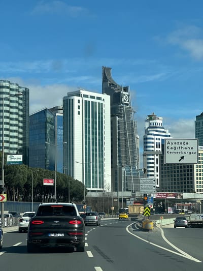 The image displays an urban roadway scene in the Maslak district of Istanbul, Turkey, under a clear blue sky with scattered white clouds. The perspective is from inside a vehicle traveling on a multi-lane asphalt road.

In the immediate foreground, a black Bentley Bentayga SUV, bearing Turkish license plate 07 BMZ 777, is positioned centrally in a lane, with its rear brake lights illuminated. Other vehicles are visible in surrounding lanes and further down the road, including a silver SUV directly behind the Bentley and a yellow car in the distance on the right. The roadway features white lane markings. To the right, a concrete barrier with yellow and black chevron markings separates the main road from an exit or merging lane.

A large white overhead road sign with black text points right to "Ayazağa Kağıthane Kemerburgaz." To the left, a dense line of coniferous trees partially obscures a sign for "British School Istanbul." The midground and background are dominated by a modern high-rise skyline. Prominent buildings include a tall, slender skyscraper with a distinctive curved upper facade, a building with a large circular corporate logo, and a white and blue-striped building displaying "OM DENTAL." Additional road and building signs are visible, indicating destinations such as "Beykent Üniversitesi" and "MEF Üniversitesi." No individuals are explicitly discernible. The scene is brightly illuminated by direct sunlight.