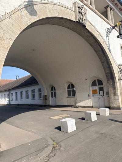 An exterior daytime photograph depicts a large, light-colored architectural archway, part of the Sprudelhof complex in Bad Nauheim, Germany. The arch features a textured, stone-like exterior and a smooth, off-white underside, framing a view of a long, single-story building with a white facade and a dark brown tiled roof, visible on the left. This building has multiple rectangular windows with white frames and a chimney on its roof.
The structure forming the archway on the right has a textured off-white wall. Beneath the arch, three arched openings are visible: a plain white door on the left, a second plain white door in the middle with a white sign on a pole beside it, and a double white door with glass panes on the right. The rightmost door displays several informational signs, including text partially visible as "Kneipp," and an intercom panel. Above the arch, a stone relief carving depicting figures is affixed to the building's facade, and an ornate black street lamp with a golden top extends from the wall further right.
The foreground consists of asphalt pavement exhibiting minor cracking and some small green plant growth near the lower left edge. Two square metal utility covers are embedded in the asphalt. Three light-colored concrete cube bollards are positioned on the pavement, casting distinct shadows towards the lower right. The sky visible through the arch is clear and blue. No individuals or vehicles are present.