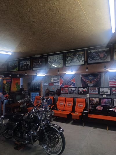 The image shows the interior of a garage or workshop, likely in Surat Thani City Municipality, Thailand, decorated with motorcycle-related memorabilia. A motorcycle is prominently displayed in the foreground. A man is sitting in an orange Harley-Davidson-themed chair. There are other orange chairs and a couch crafted from oil drums, also Harley-Davidson-themed. Walls are covered in corrugated metal and decorated with framed motorcycle posters, flags (including a Confederate flag and an American flag), and license plates. A vintage television and other items are placed on a wooden table in the background.