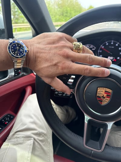 The image captures a close-up, first-person perspective from inside a luxury vehicle, likely a Porsche, located in Ingeldorf, Luxembourg. The central focus is a person's right hand holding the steering wheel, adorned with a distinctive watch and ring.
The person's hand, with tan skin and neatly trimmed nails, grips the black leather steering wheel. The steering wheel features a prominent Porsche crest in its center, displaying the "PORSCHE" text above a golden shield with red and black stripes and a black horse. Partially visible behind the steering wheel are the car's gauges, with a digital readout showing "86", possibly indicating speed.
On the ring finger of the right hand, a large, ornate gold ring with intricate, possibly serpentine, designs is worn. On the left wrist, a two-tone (steel and gold) Rolex Submariner watch is visible, featuring a vibrant blue dial and bezel with gold markings. Text such as "ROLEX", "OYSTER PERPETUAL", "SUBMARINER", "1000ft=300m", and "SUPERLATIVE CHRONOMETER OFFICIALLY CERTIFIED" can be seen on the watch face.
The car's interior features rich red leather accents on the door panel or console, with various control buttons also visible. The person's leg is dressed in light-colored (beige or khaki) cargo-style pants. Bright daylight streams in from outside the car, suggesting it is daytime. The overall scene conveys a sense of luxury and travel.