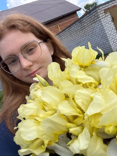 The image presents a close-up, rotated portrait of a young person outdoors, holding or positioned next to a large bouquet of pale yellow irises. The individual has fair skin with scattered freckles visible on their nose and cheeks, and long, reddish-brown hair. They wear thin, square-framed glasses with light-colored rims, through which their light blue-green eyes are visible. Their lips are slightly parted, and their expression appears neutral or contemplative.
A prominent feature is the large cluster of yellow irises, which dominates the upper right side of the frame. The flowers are a soft, delicate yellow, with some showing subtle striped patterns on their inner petals. The person's dark clothing is partially visible in the lower right corner.
The background provides glimpses of an outdoor setting. Behind the person, structures with a dark, possibly corrugated roof and brown siding are visible, alongside a patch of blue sky with white clouds and some green tree foliage. Behind the flowers, a light-colored, possibly brick or paved, surface can be seen. The overall lighting suggests it is daytime. No visible text is present. The scene captures a tranquil moment in what appears to be a residential or garden environment.