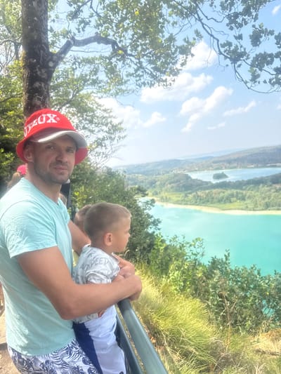 In the city of La Chaux-du-Dombief, France, a man wearing a red bucket hat with "LUX" printed on it and a light blue t-shirt is standing next to a young boy. The man has his arm around the boy. The boy is wearing a light-colored t-shirt with a print and blue and white shorts. They are both looking out at a scenic view of lakes and hills. The environment is outdoors with trees and a bright sky. The image appears to be taken on a sunny day, likely during the daytime.